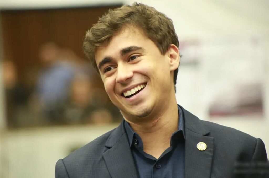 Nikolas Ferreira sorri durante evento público usando terno escuro.