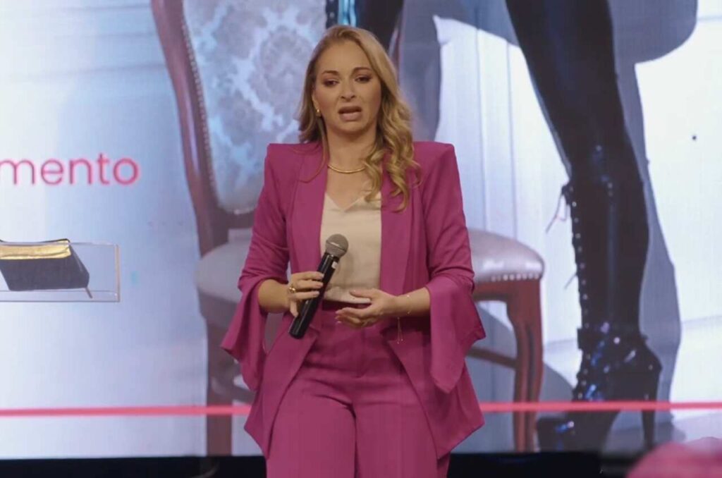 Pastora Patrícia Fernandes ministra palestra em evento cristão, segurando microfone e falando ao público em palco iluminado.