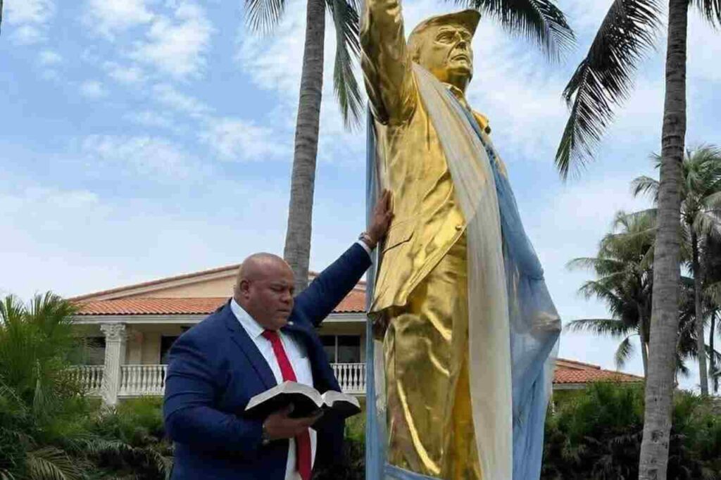 Pastor Mark Burns ao lado de estátua dourada de Donald Trump segurando Bíblia em área externa.