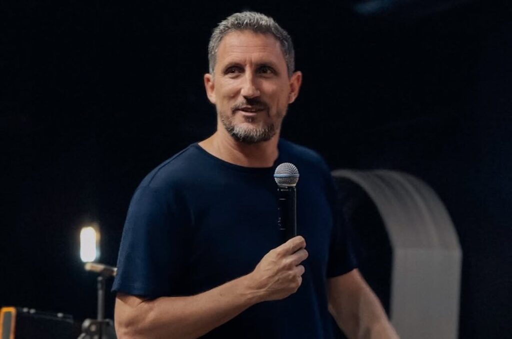 Pastor Telmo Martinello segura um microfone durante ministração em ambiente de igreja, diante de fundo escuro.