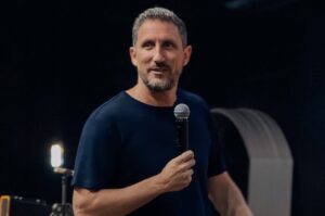 Pastor Telmo Martinello segura um microfone durante ministração em ambiente de igreja, diante de fundo escuro.