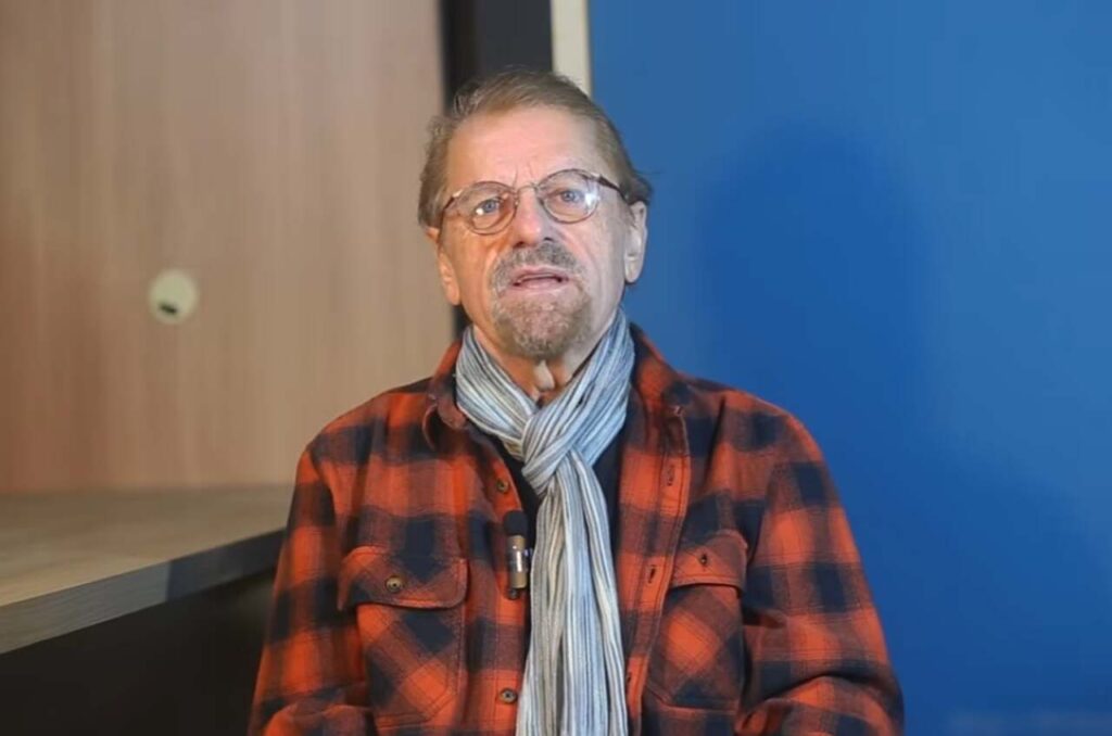 O cantor Asafe Borba aparece sentado durante uma gravação em vídeo, usando camisa xadrez vermelha, cachecol cinza e óculos.