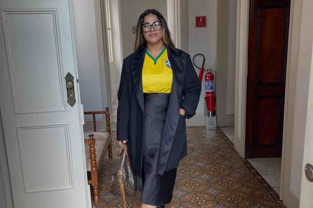 Michelle Nascimento posa em corredor de prédio histórico usando camiseta da seleção brasileira, sobretudo preto e óculos.