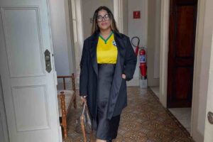 Michelle Nascimento posa em corredor de prédio histórico usando camiseta da seleção brasileira, sobretudo preto e óculos.