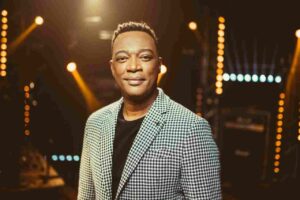 Marquinhos Gomes posa para foto em palco iluminado, usando blazer xadrez e camiseta preta durante evento gospel.
