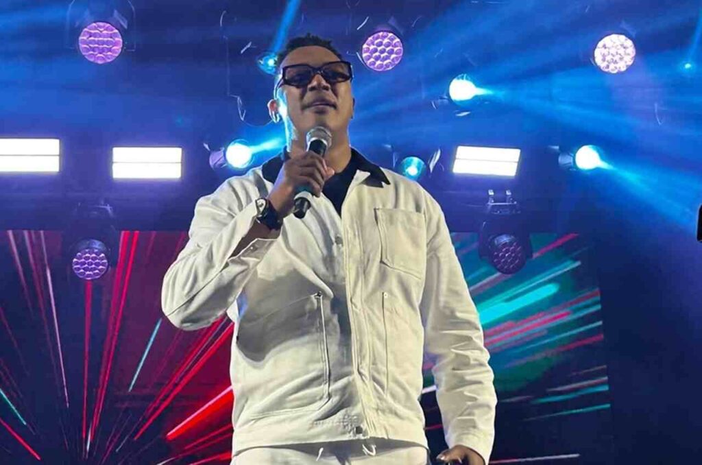 Marquinhos Gomes canta em apresentação ao vivo segurando um microfone diante de palco iluminado com efeitos visuais coloridos.