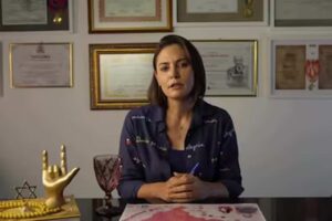 Michelle Bolsonaro aparece sentada à mesa em um escritório, gravando um pronunciamento diante de uma parede com certificados e homenagens emoldurados.