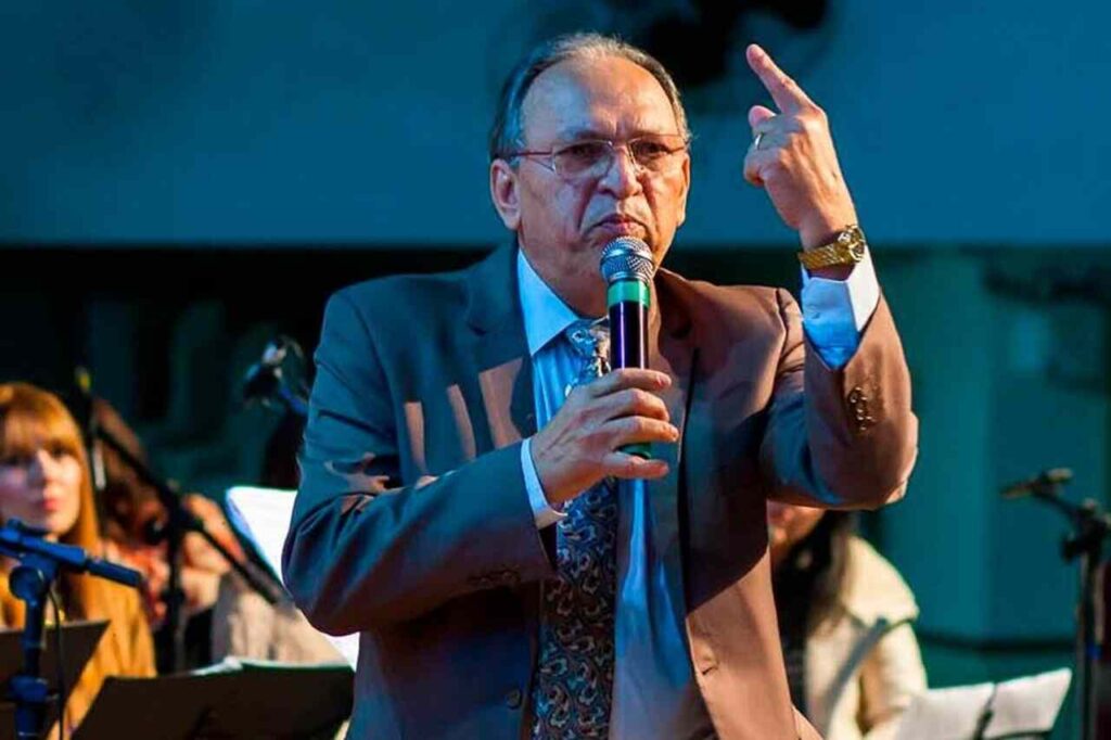 Pastor Napoleão Falcão prega ao microfone durante culto, gesticulando diante da congregação em evento cristão.