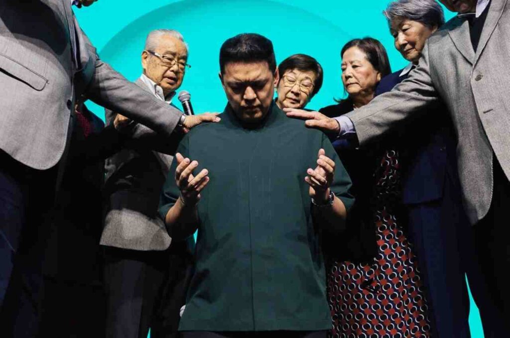Pastor Téo Hayashi recebe oração com imposição de mãos durante cerimônia religiosa realizada diante de uma congregação.