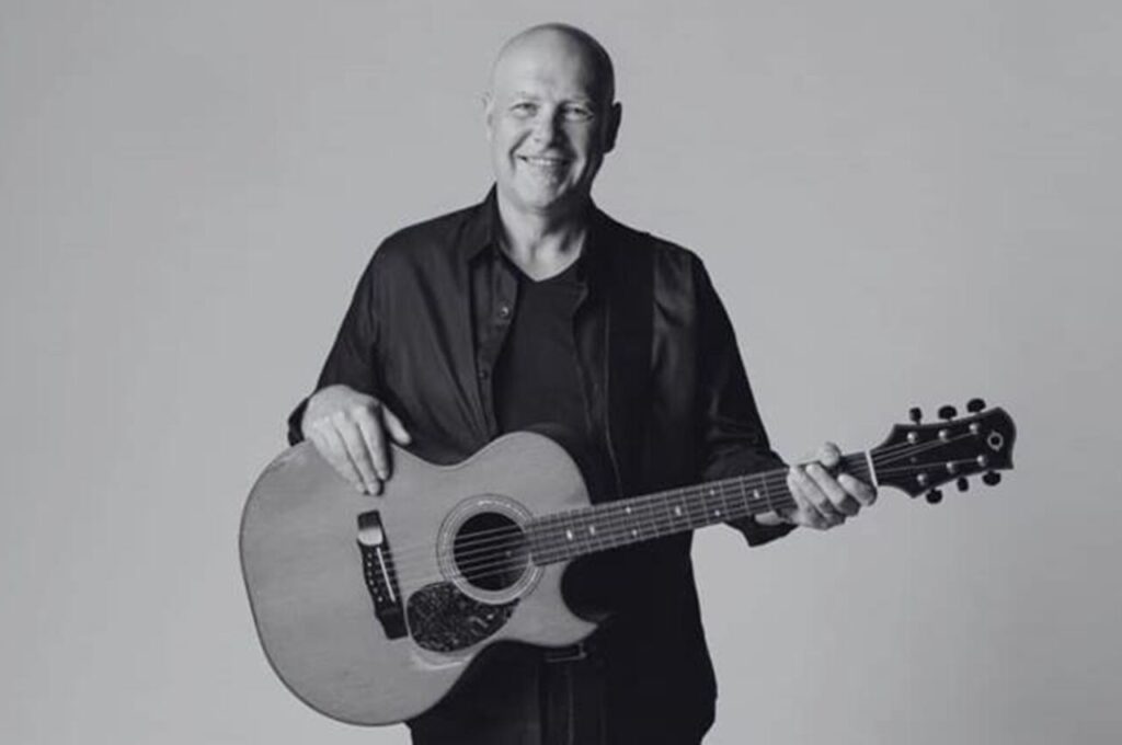 O cantor Brian Doerksen posa sorrindo enquanto segura um violão em um retrato em preto e branco.