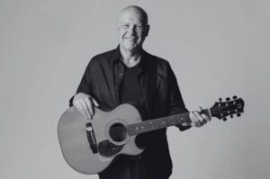 O cantor Brian Doerksen posa sorrindo enquanto segura um violão em um retrato em preto e branco.