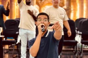 Cantor João Marcos ministra louvor com um microfone nas mãos, apontando para o público durante culto em igreja.
