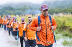 Integrantes do movimento Legendários caminham em fila durante uma trilha ao ar livre, usando mochilas e uniformes laranja.