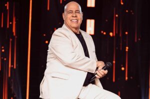 O cantor gospel Mattos Nascimento aparece sorrindo no palco durante uma apresentação, segurando um microfone e usando um terno claro.