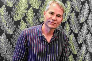 Pastor Márcio Poncio posa para fotografia usando camisa listrada diante de um painel com estampa de folhas.