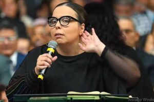 A pastora Helena Raquel ministra durante um culto, segurando um microfone e uma Bíblia diante da igreja.