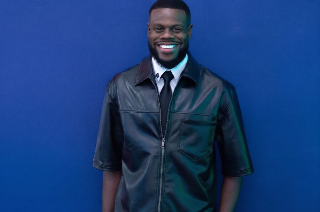 Cantor Samuel Eleotério posa sorrindo diante de um fundo azul, usando jaqueta preta de couro, camisa branca e gravata.