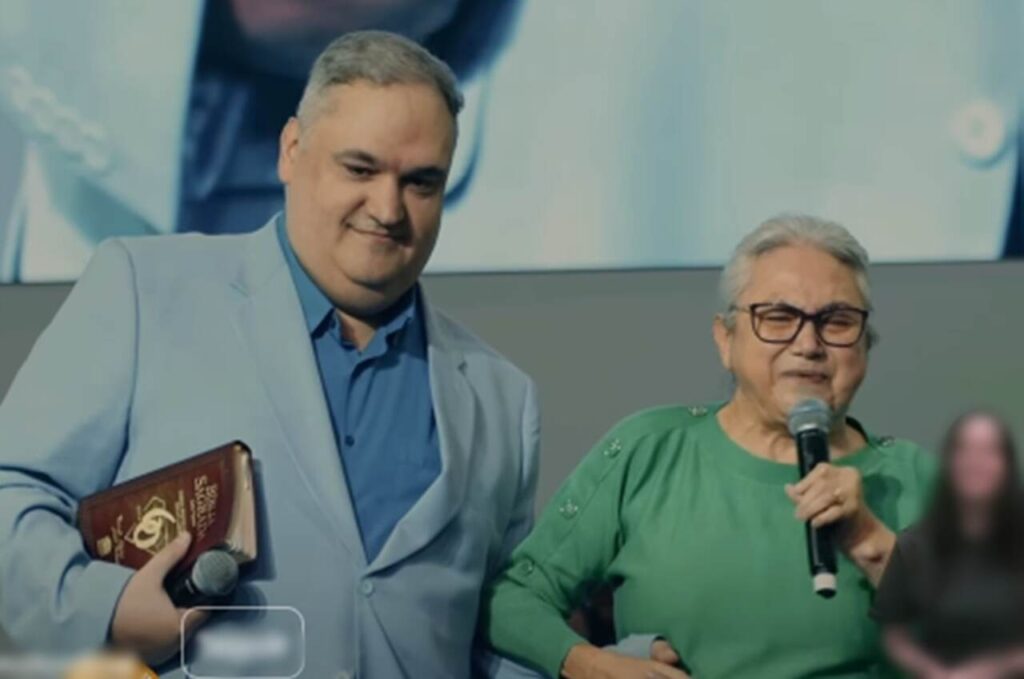 David Miranda Filho aparece ao lado da mãe durante evento religioso; ela segura um microfone enquanto ele segura uma Bíblia.