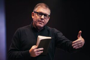 Pastor Ed René Kivitz prega segurando uma Bíblia e gesticulando durante mensagem em conferência cristã.