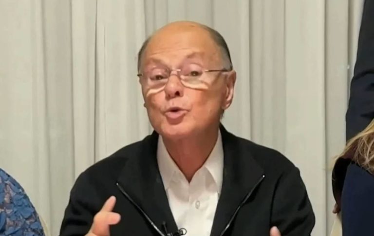 Edir Macedo fala durante entrevista, usando óculos e camisa branca com jaqueta preta.
