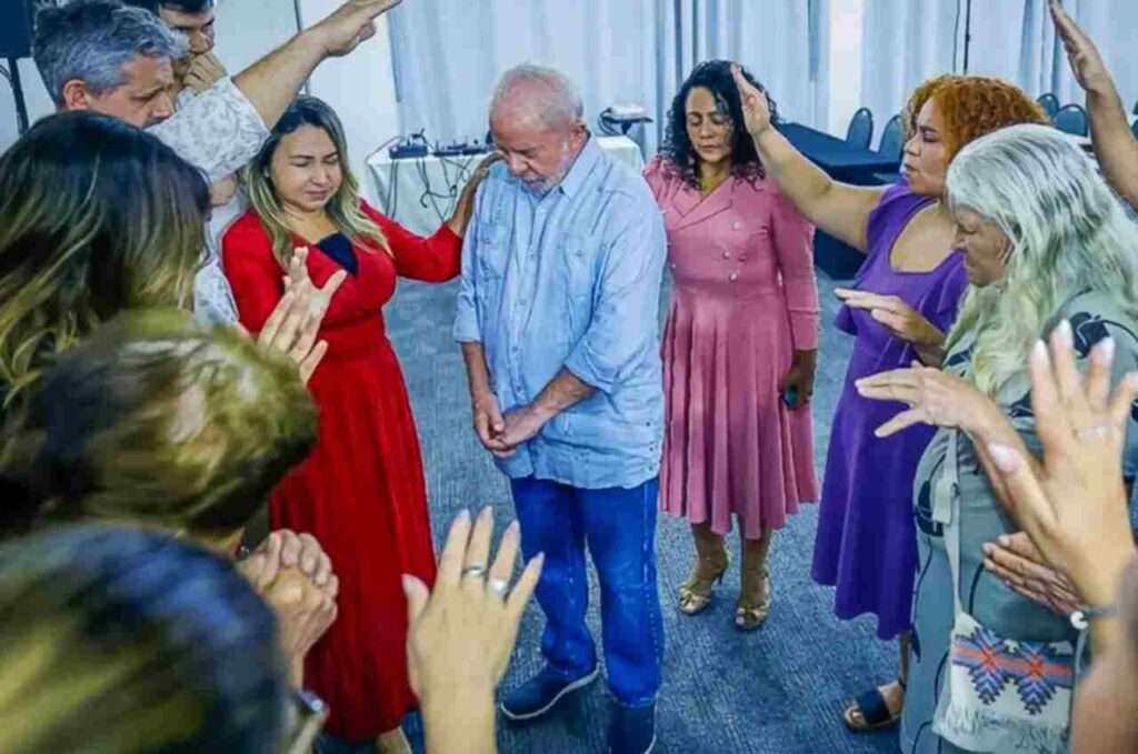 Homem de cabelos brancos participa de um momento de oração cercado por pessoas com as mãos levantadas.