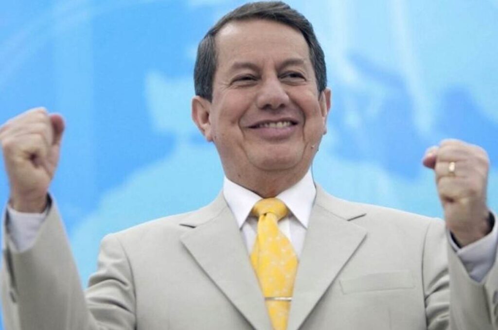 Missionário R. R. Soares aparece sorrindo, usando terno claro e gravata amarela, com os braços levantados.
