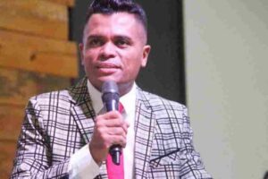 Pastor João Caetano prega segurando um microfone durante culto em igreja evangélica, usando terno xadrez e gravata vermelha.