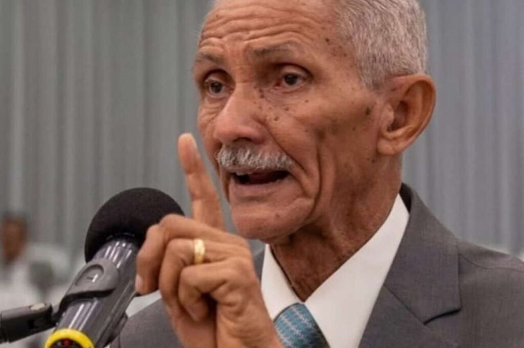 : Pastor José Carlos de Lima prega ao microfone com o dedo indicador levantado durante culto em igreja evangélica.