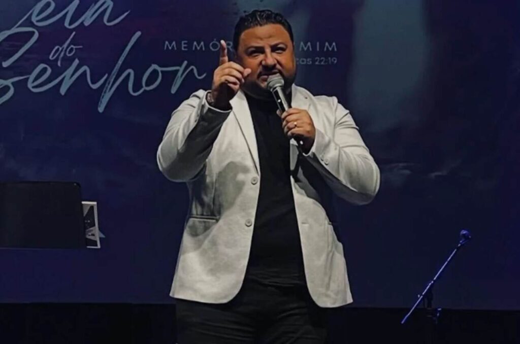 Pastor Moisés de Jesus ministra ao microfone durante culto, usando terno branco e camisa preta.