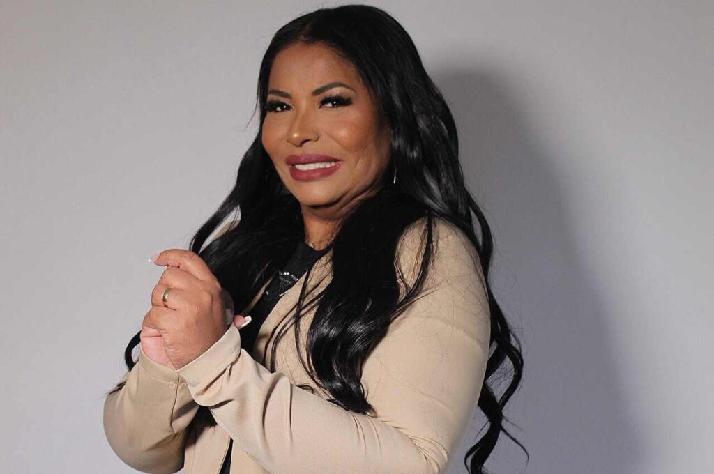 Pastora Elizângela Terra aparece sorrindo, com cabelos longos e pretos, usando blazer bege e blusa preta.