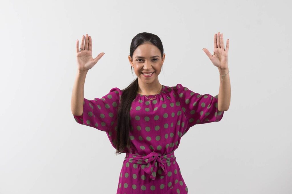 Cantora gospel Vitória Souza sorri com as mãos levantadas durante ensaio fotográfico promocional em fundo branco.