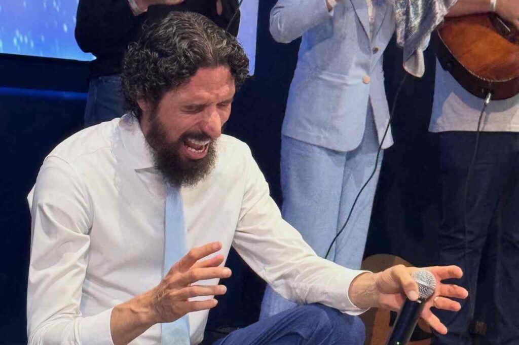 Apóstolo Robson Martins aparece ajoelhado durante um momento de oração, segurando um microfone e acompanhado por músicos.