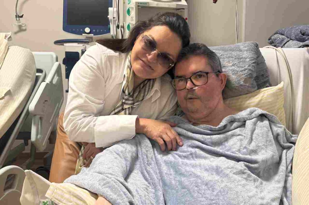 Mulher abraça o marido internado em quarto de hospital enquanto os dois posam para uma foto