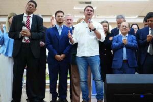 Homem de camisa branca fala ao microfone durante culto evangélico ao lado de pastores e líderes religiosos