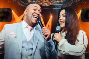 Gerson Rufino e Antônia Gomes cantam juntos durante apresentação musical gospel, segurando microfones sob iluminação cênica alaranjada.