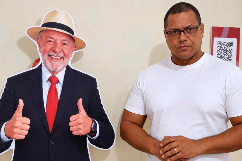 Homem posa ao lado de um recorte de papelão do presidente Lula sorrindo, usando chapéu, terno preto e gravata vermelha.