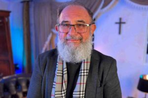 Pastor Alex Alves usa óculos, blazer cinza e cachecol xadrez em retrato feito dentro de uma igreja, com uma cruz ao fundo.