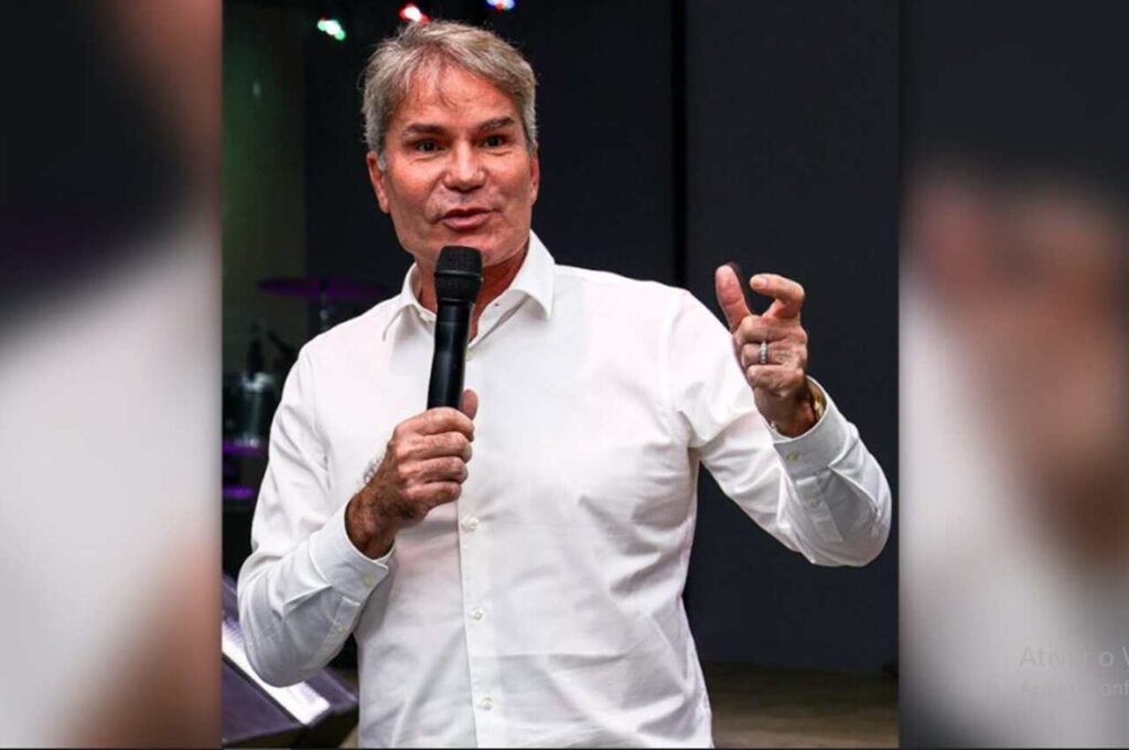 Pastor Márcio Poncio segura um microfone e faz um gesto com a mão durante uma pregação ou palestra, usando camisa social branca.