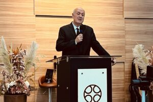 Pastor Renato Vargens, de terno preto e gravata, segura um microfone e ministra atrás de um púlpito em uma igreja com paredes de madeira.