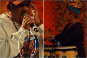 Pregador Luo canta ao microfone usando colar religioso, enquanto percussionista toca atabaques em apresentação musical.