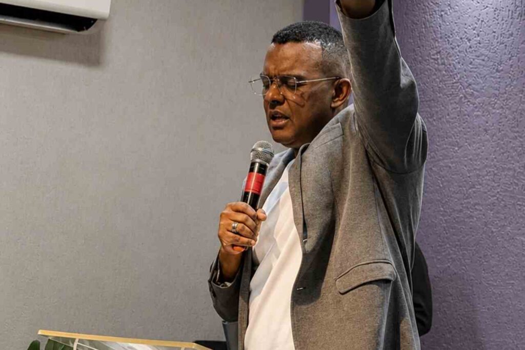 Renato de Paula ministra em igreja com microfone na mão e um dos braços levantados, usando terno cinza e óculos.