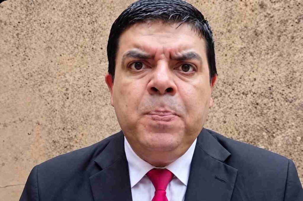 Homem de terno preto, camisa branca e gravata vermelha posa para fotografia com expressão séria diante de uma parede de textura bege