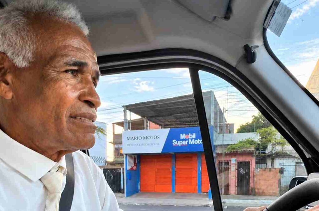Sargento Isidório aparece sentado ao volante de um carro, usando camisa branca e gravata clara, enquanto dirige por uma rua urbana.