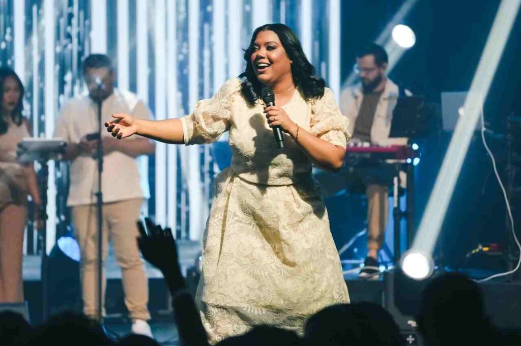 A cantora gospel Sued Silva se apresenta no palco com um microfone na mão, usando um conjunto bege de renda, enquanto músicos e iluminação azul compõem o cenário.